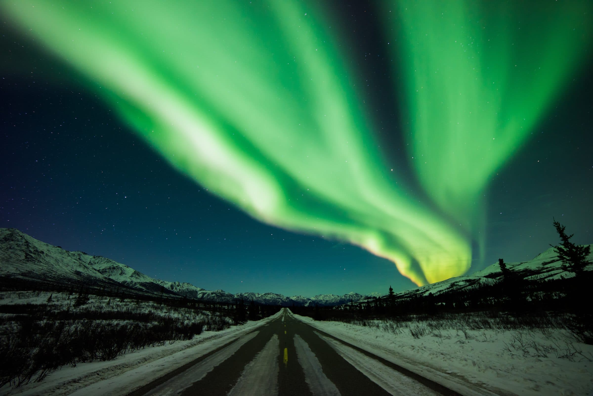 Green northern lights sweeping over a snowy road and mountains in Denali National Park
